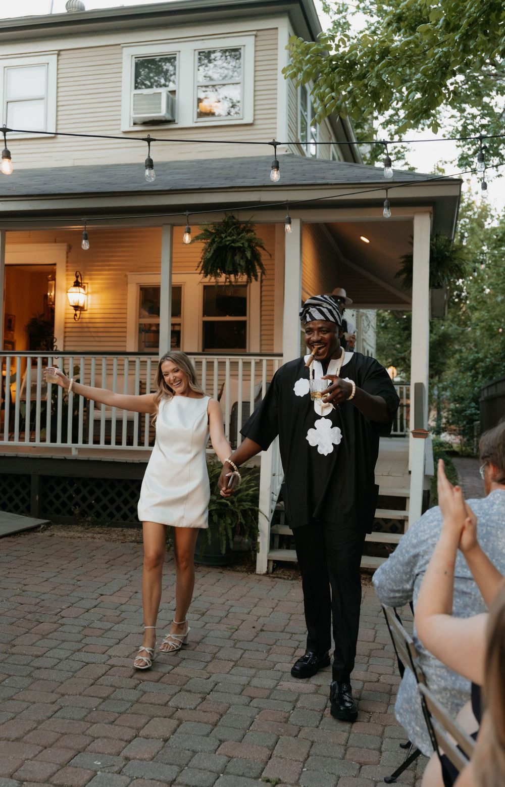 Couple celebrating outdoors in front of a charming porch with string lights at a backyard wedding reception.