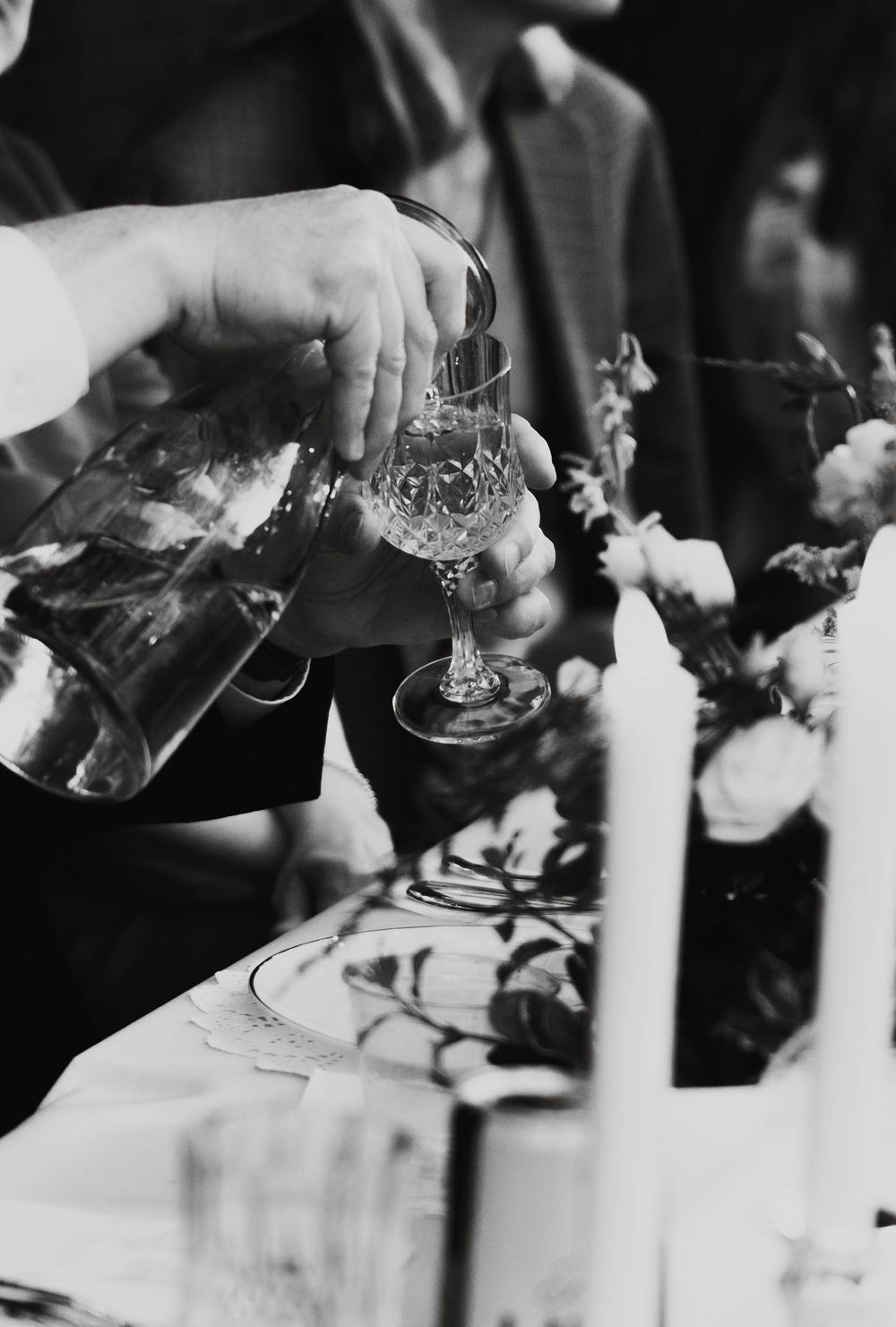 Black and white photo of a person pouring liquid into a crystal glass at an elegant floral table setting.