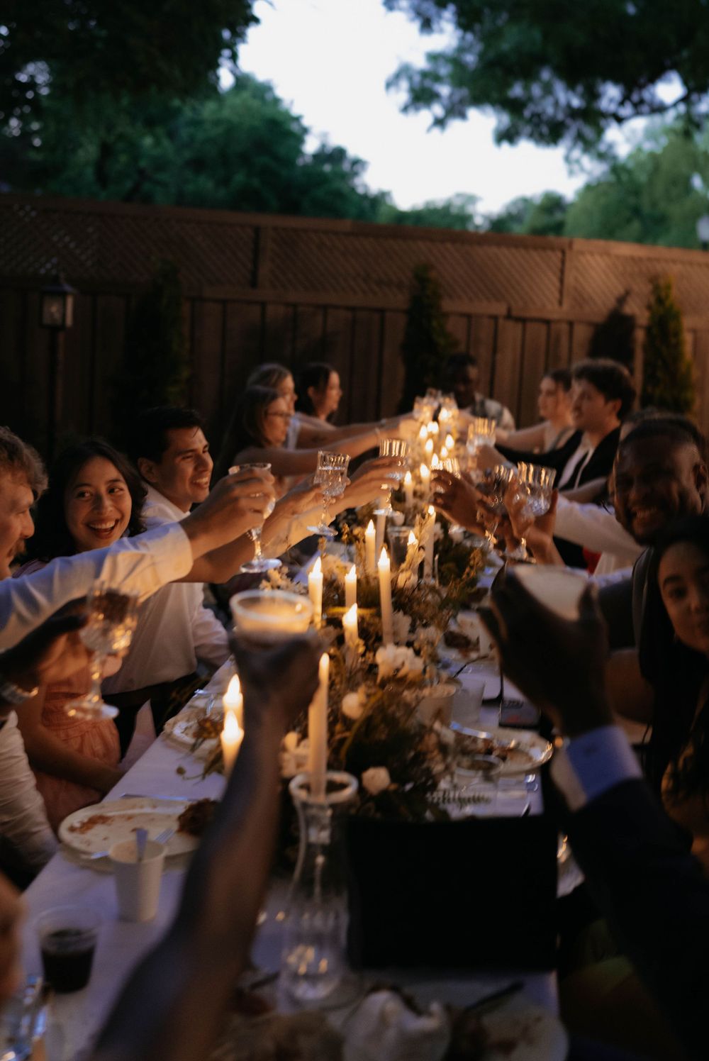 Guests raising champagne glasses in a candlelit outdoor dinner toast at an elegant evening wedding reception.
