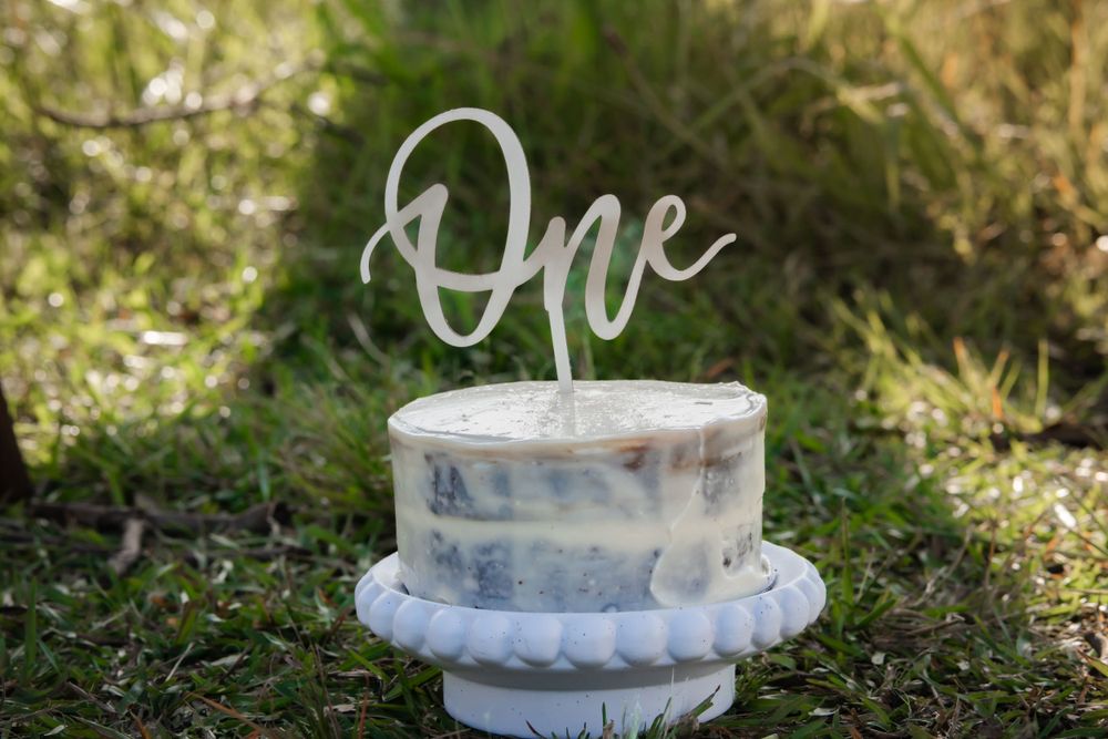 A small white birthday cake with a silver 'One' topper sits on a white pedestal stand in outdoor grass setting.