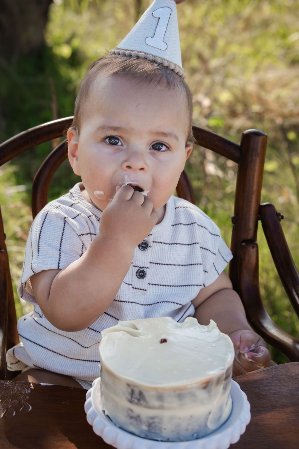 Baby wearing number 1 party hat tastes birthday cake while sitting in wooden chair at outdoor celebration.