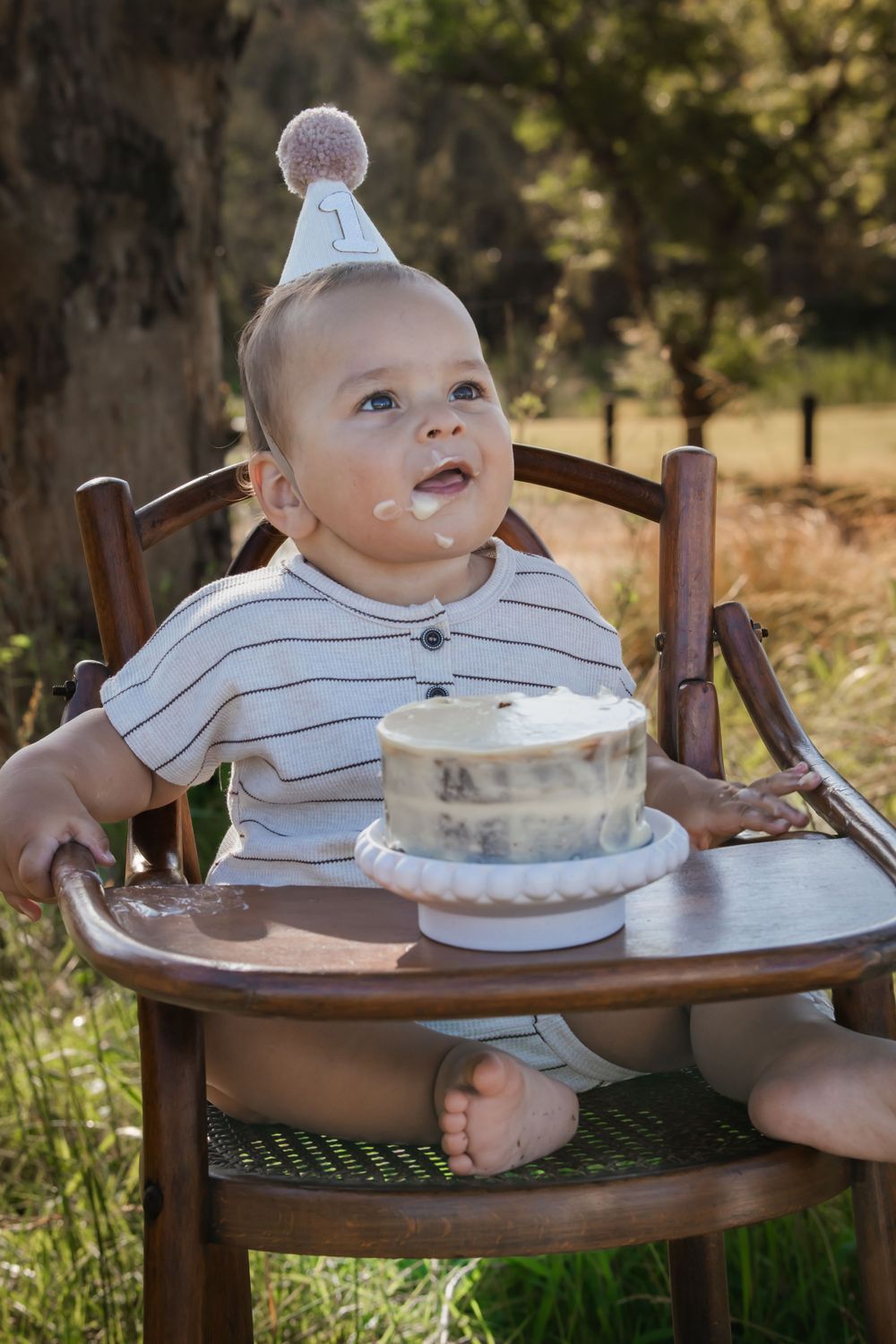 Small child in party hat sits in wooden highchair with smash cake and frosting on face during outdoor photo session.