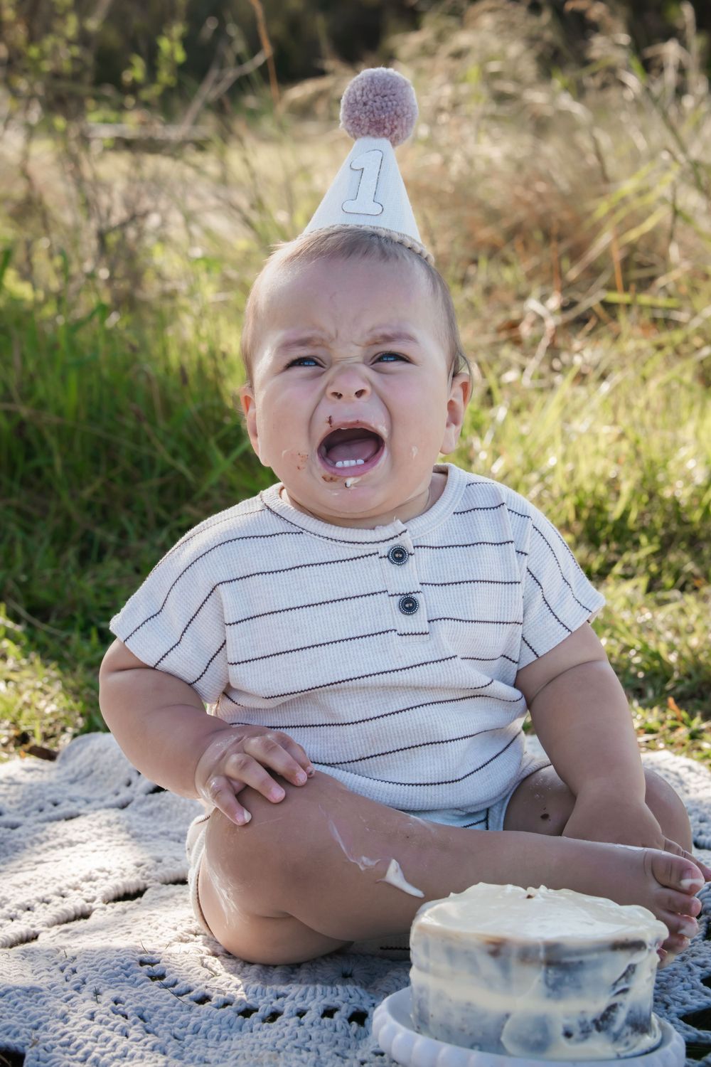 A joyful baby wearing a striped shirt and party hat sits on a blanket next to a small cake in an outdoor setting.