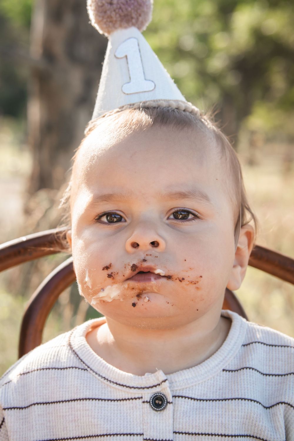 Small child wearing a birthday hat and striped shirt has chocolate cake smeared around their mouth.