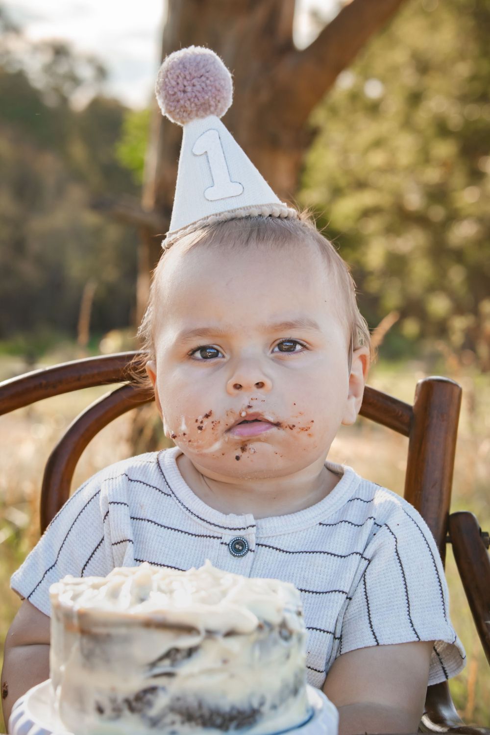 First birthday celebration with white cake and a party hat shows messy frosting around little one's mouth.