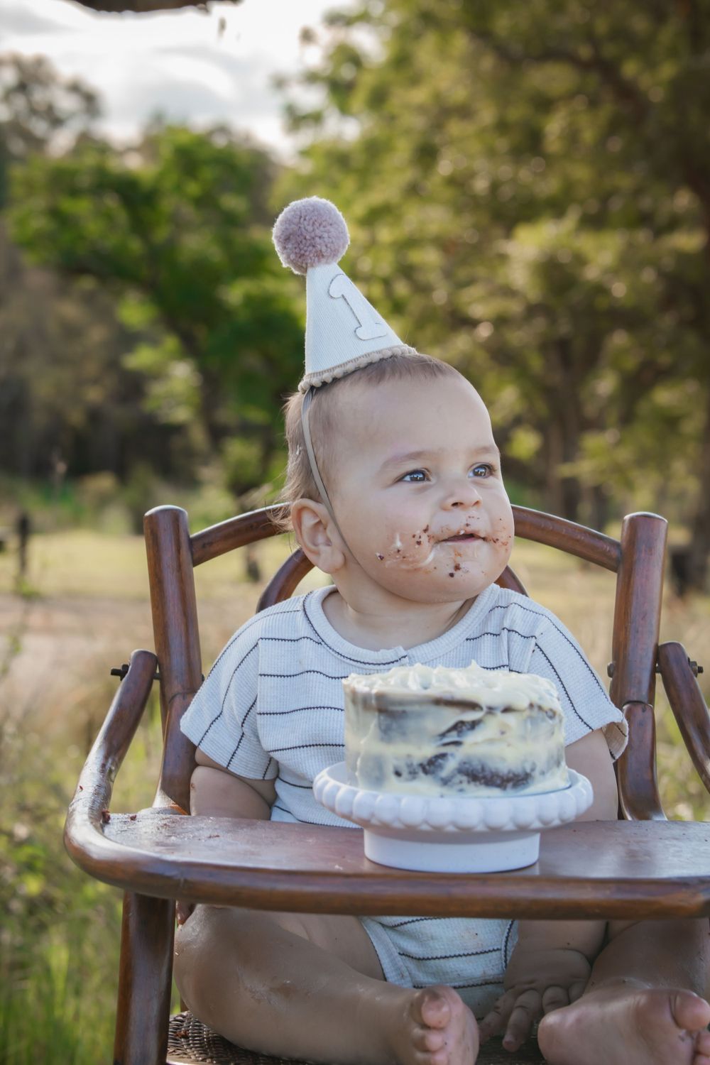 A small celebrant in a party hat sits in a wooden high chair with a smash cake dessert outdoors.
