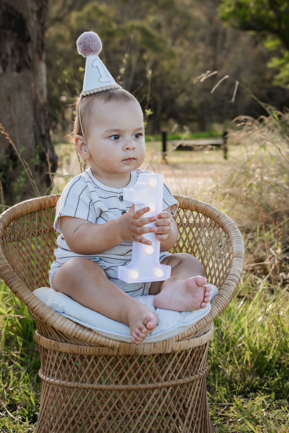 Small child wearing a party hat sits in wicker chair outdoors holding a milk bottle in a natural setting.