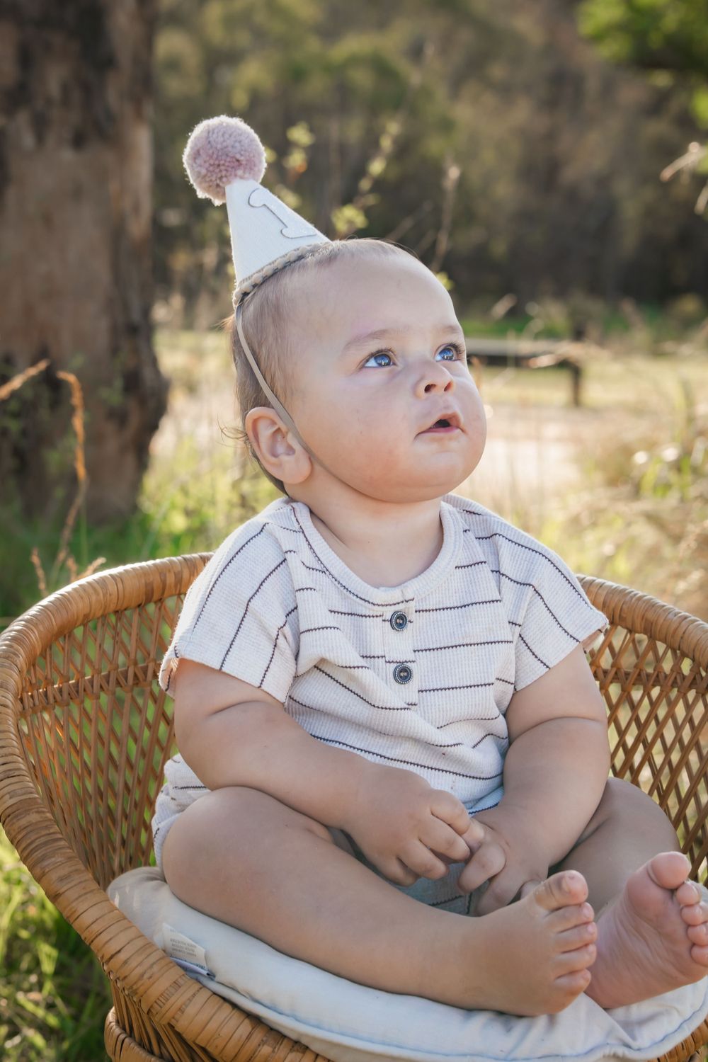Baby wearing white striped onesie and party hat sits in wicker chair outdoors during golden hour.