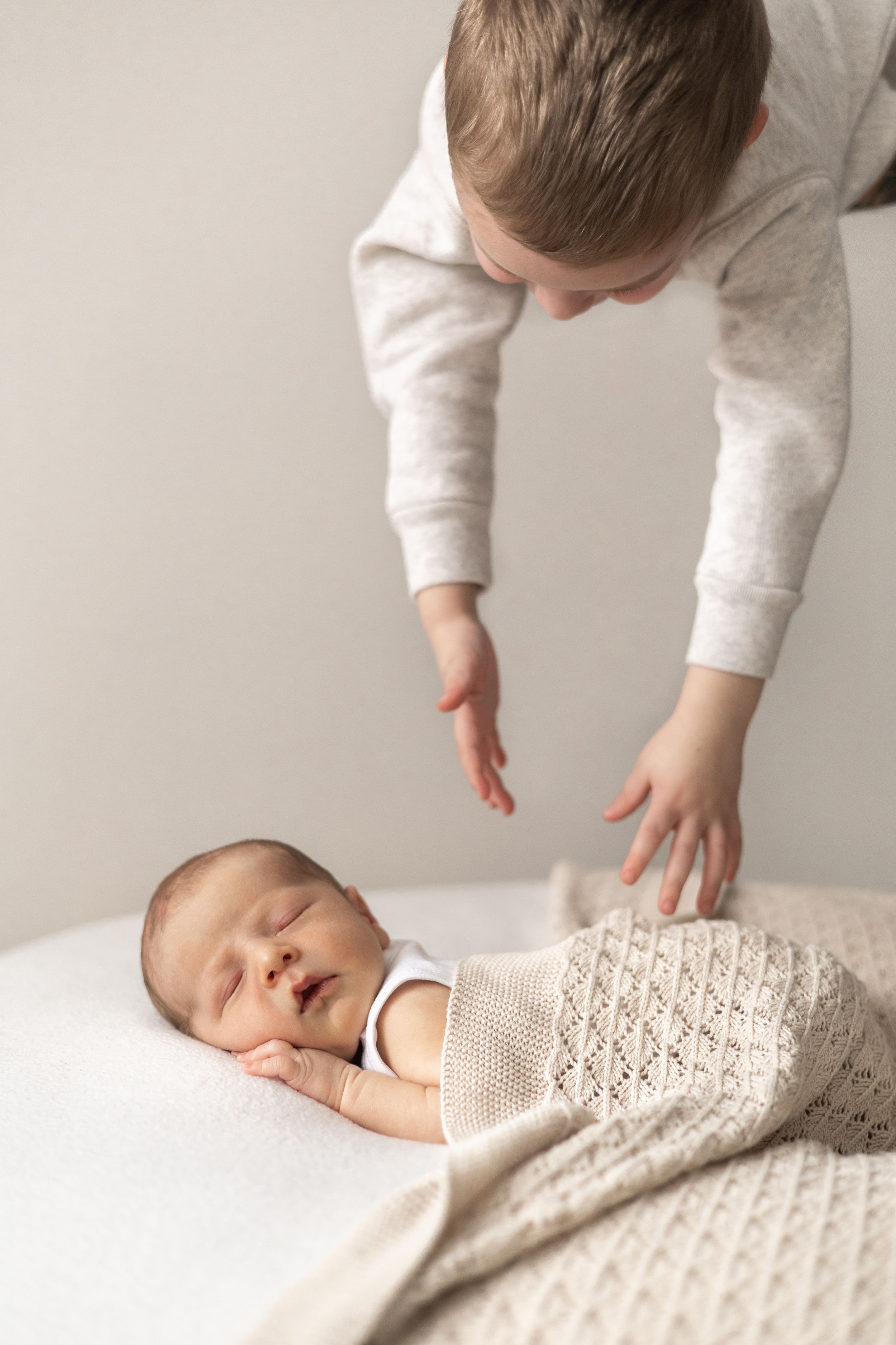 Older boy leaning over sleeping newborn baby wrapped in a cream knit blanket on a white surface.