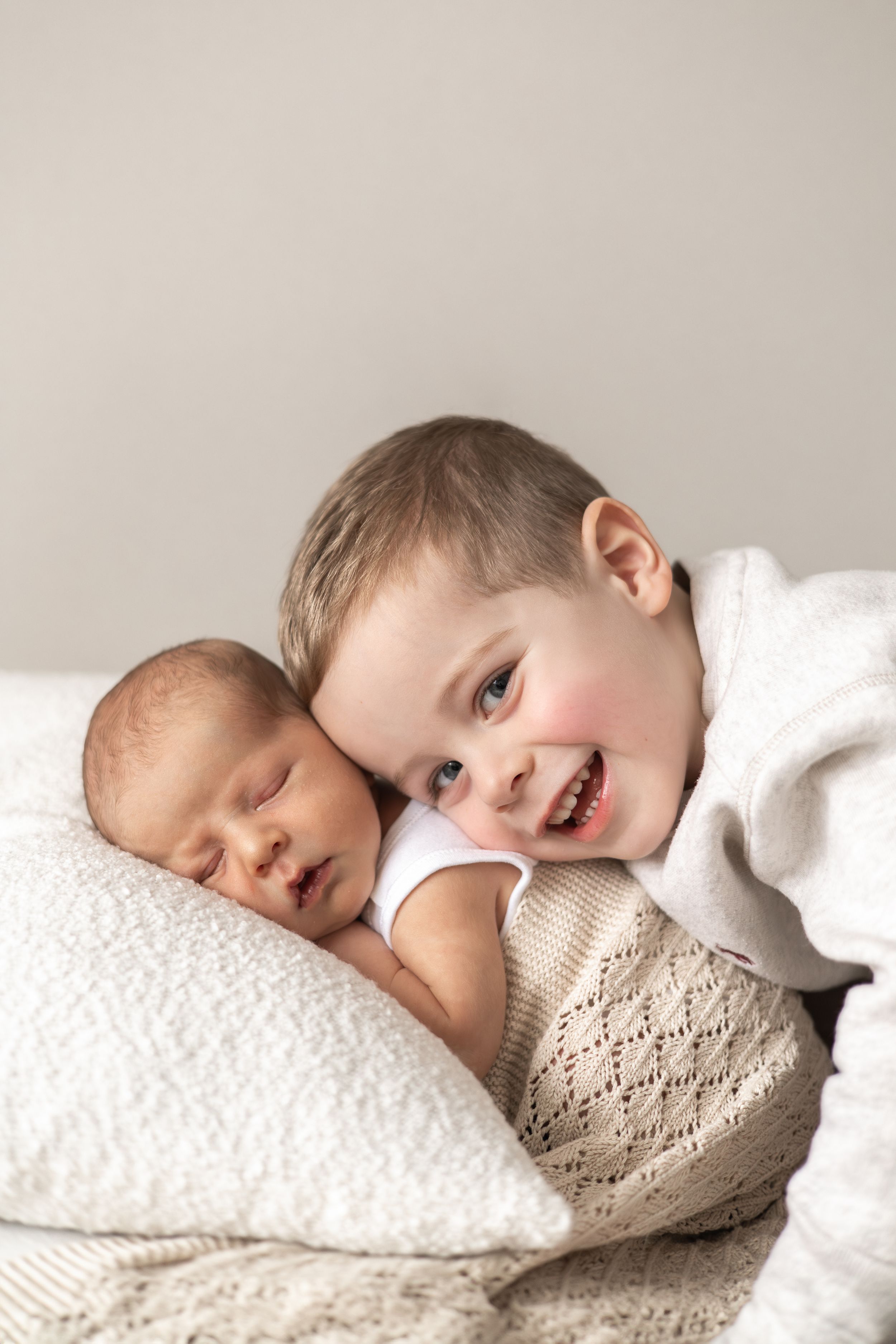 Smiling toddler gently resting cheek against sleeping newborn baby sibling on white pillow and blanket Nottingham newborn photographer