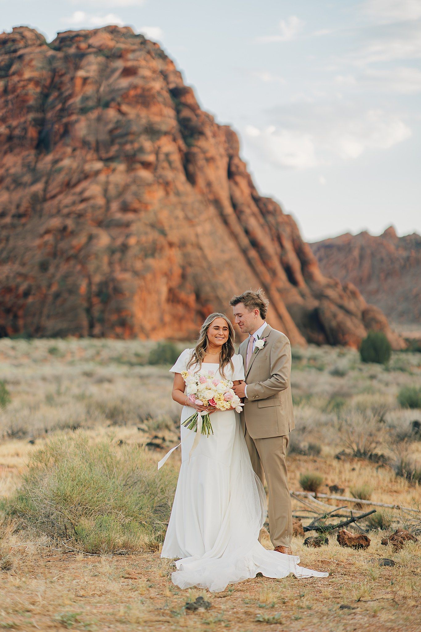 Bride and groom pose before red rock canyon landscape, she holds pastel bouquet in white gown, he wears tan suit.