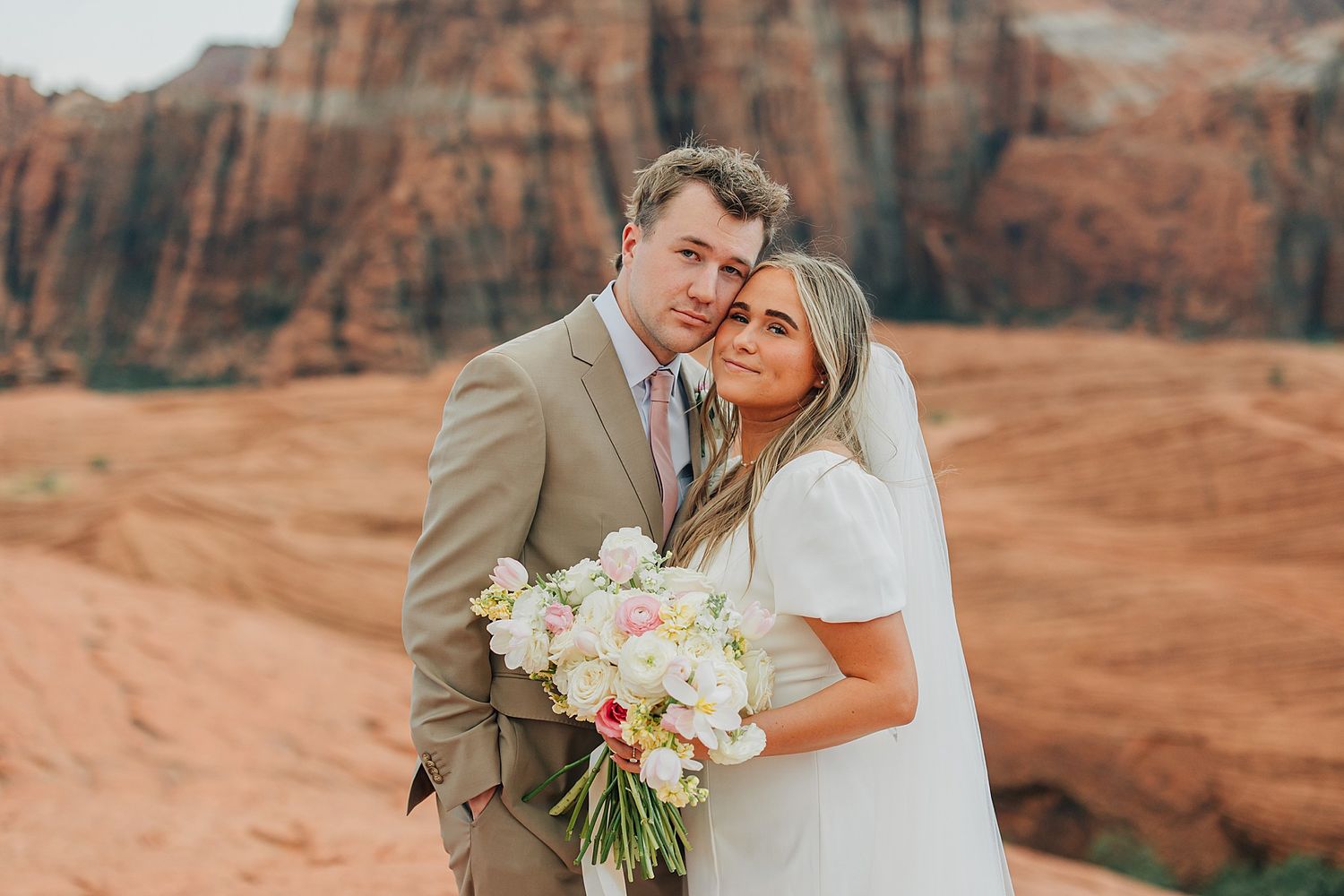 Bride and groom pose together holding a white floral bouquet with red rock canyon backdrop.