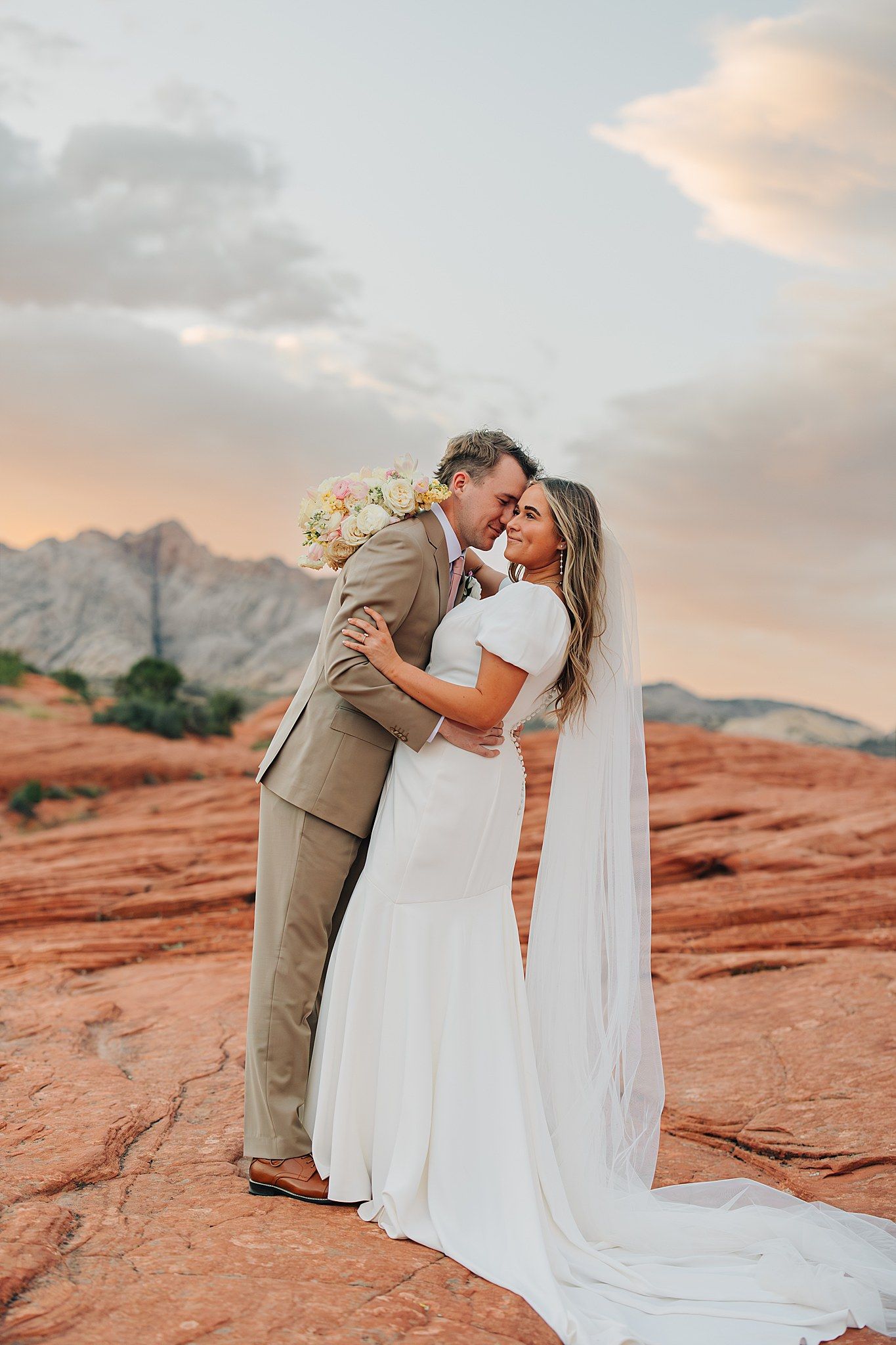 Bride and groom embrace romantically on red rock desert landscape with mountain backdrop at sunset.