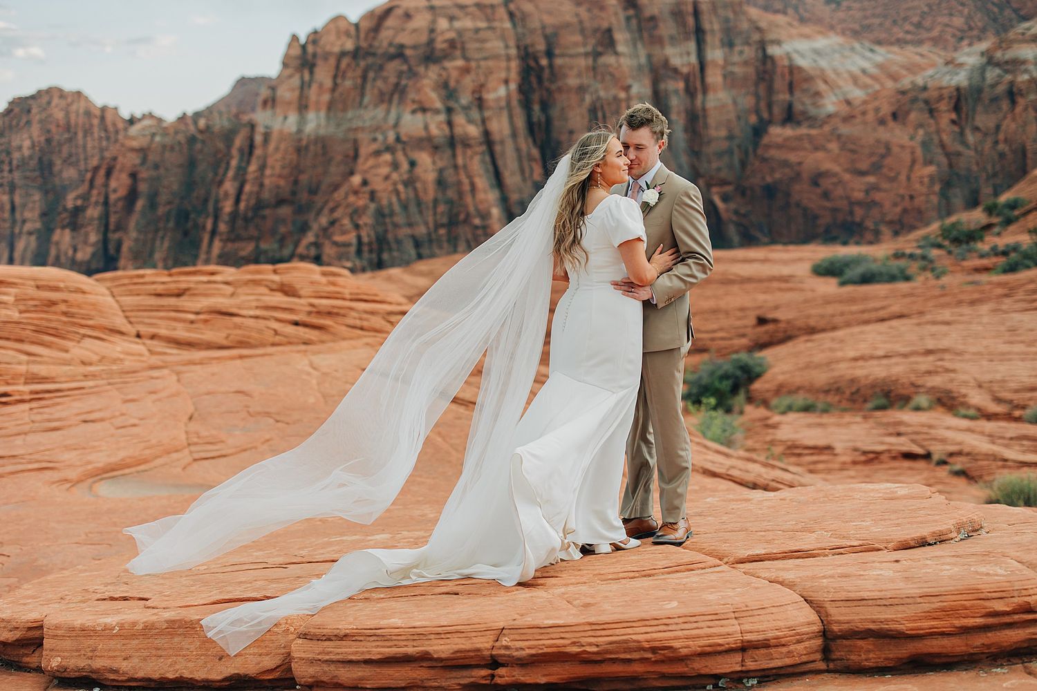 Bride in white gown with flowing veil embraces groom on red sandstone rocks with canyon backdrop.
