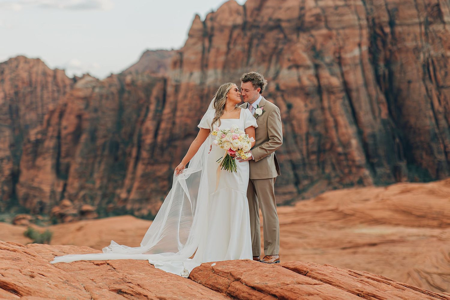 Bride and groom share a romantic kiss on red rock formations at Zion National Park with stunning canyon cliffs backdrop.
