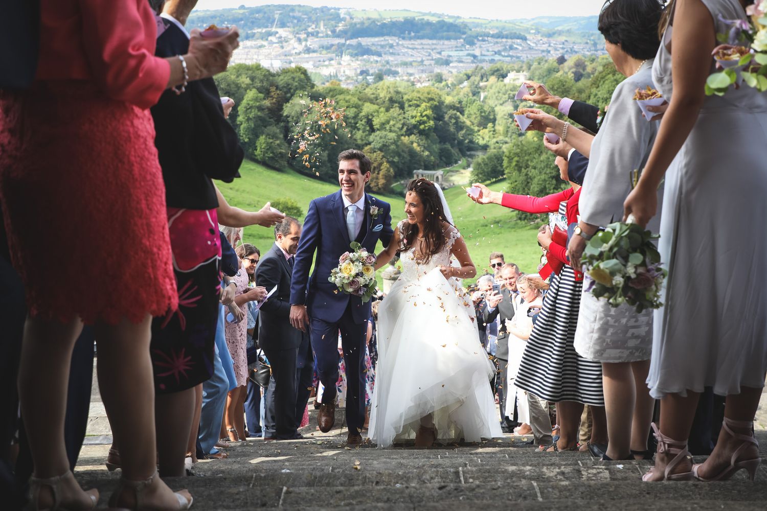 Newlywed couple walks through confetti shower on stone steps surrounded by cheering wedding guests outdoors at Prior Park Bath