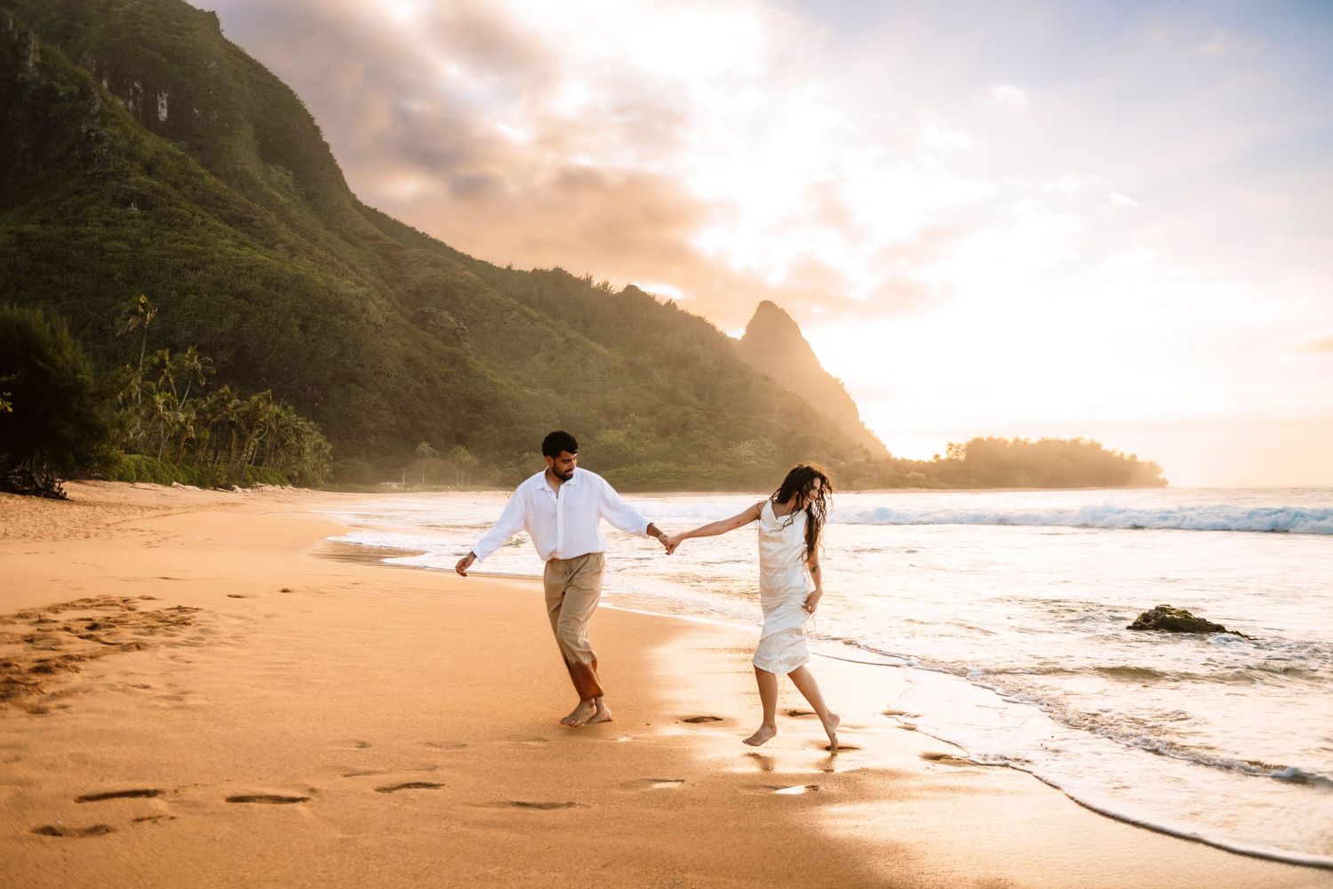 Couple holding hands walking on a golden Hawaiian beach at sunset with lush green mountains in the background.