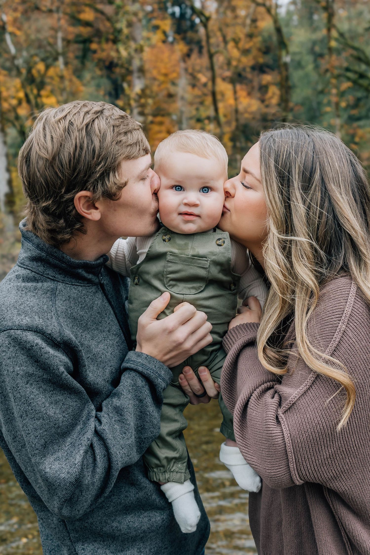 Two parents kissing their blue-eyed baby on the cheeks in an autumn forest setting.