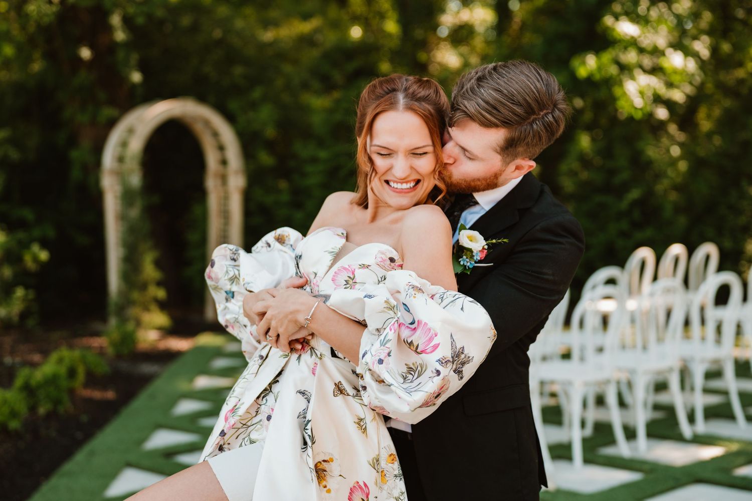 A joyful couple embraces outdoors at a wedding, bride in floral gown, groom in black tux, with white chairs and arch behind.
