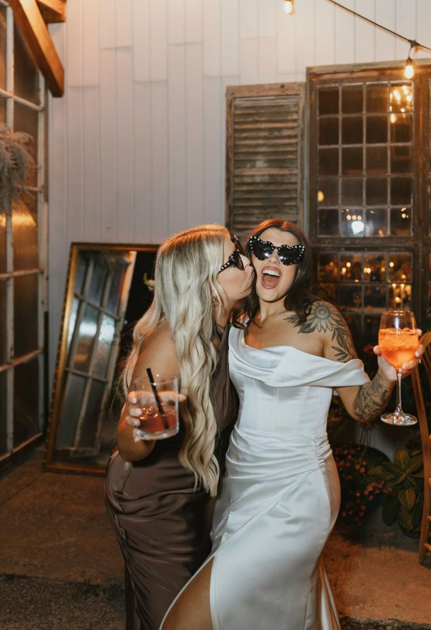 Two women in elegant dresses celebrate at a wedding, one in white with sunglasses holding a cocktail.
