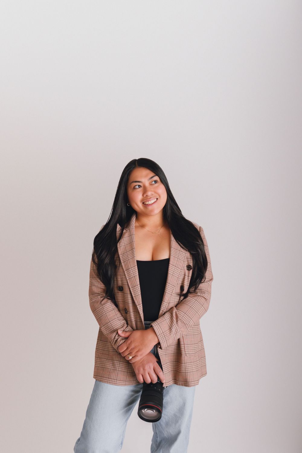 Young woman in plaid blazer holding a camera, smiling confidently against a light gray background.