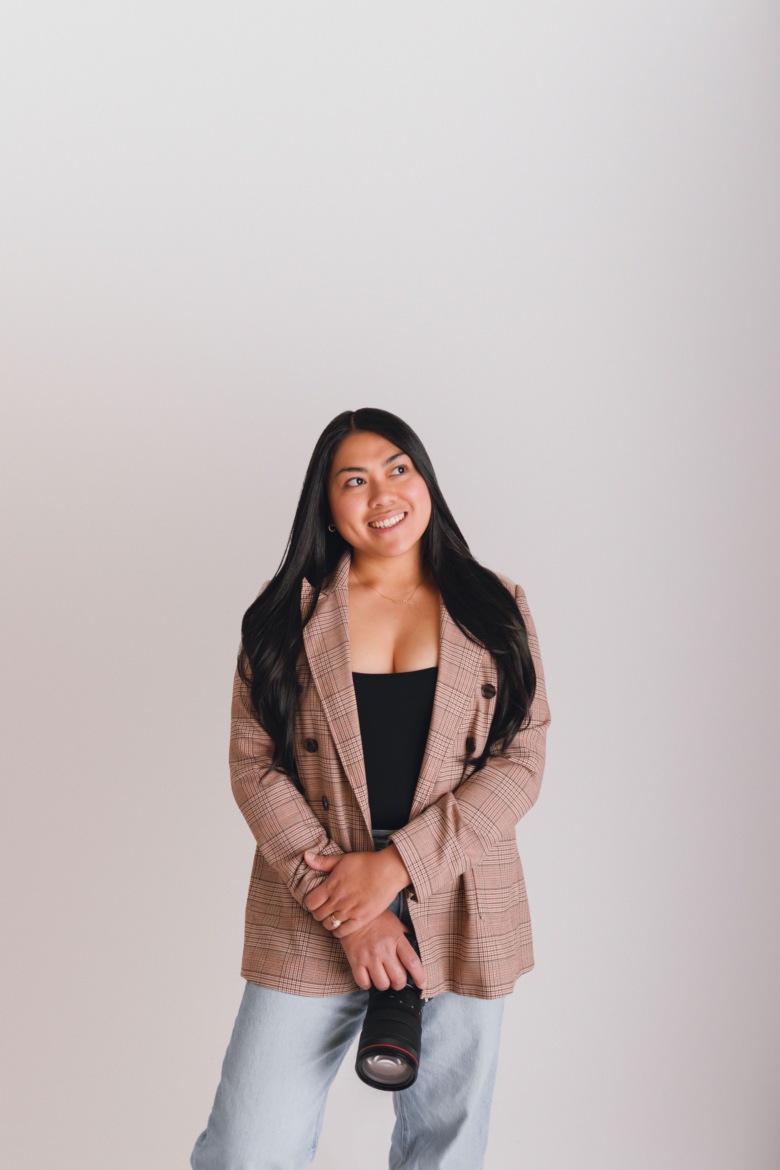 Young woman in plaid blazer holding a camera, smiling confidently against a light gray background.