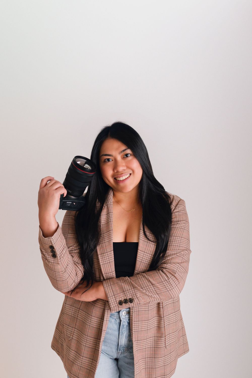 Smiling woman in plaid blazer holding a camera lens against a white background.