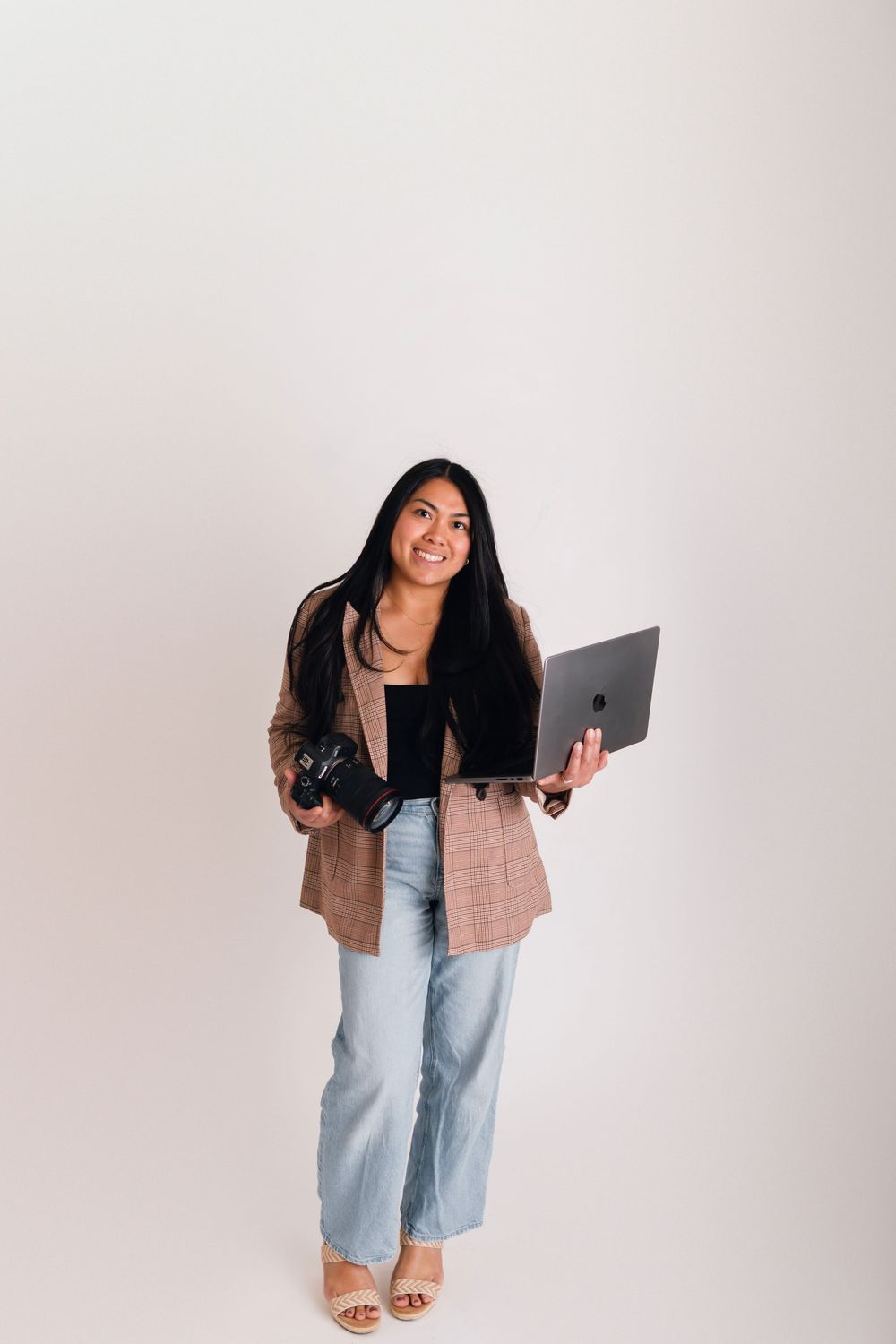 Smiling woman holding a camera and laptop, wearing a blazer and jeans, standing against a white background.