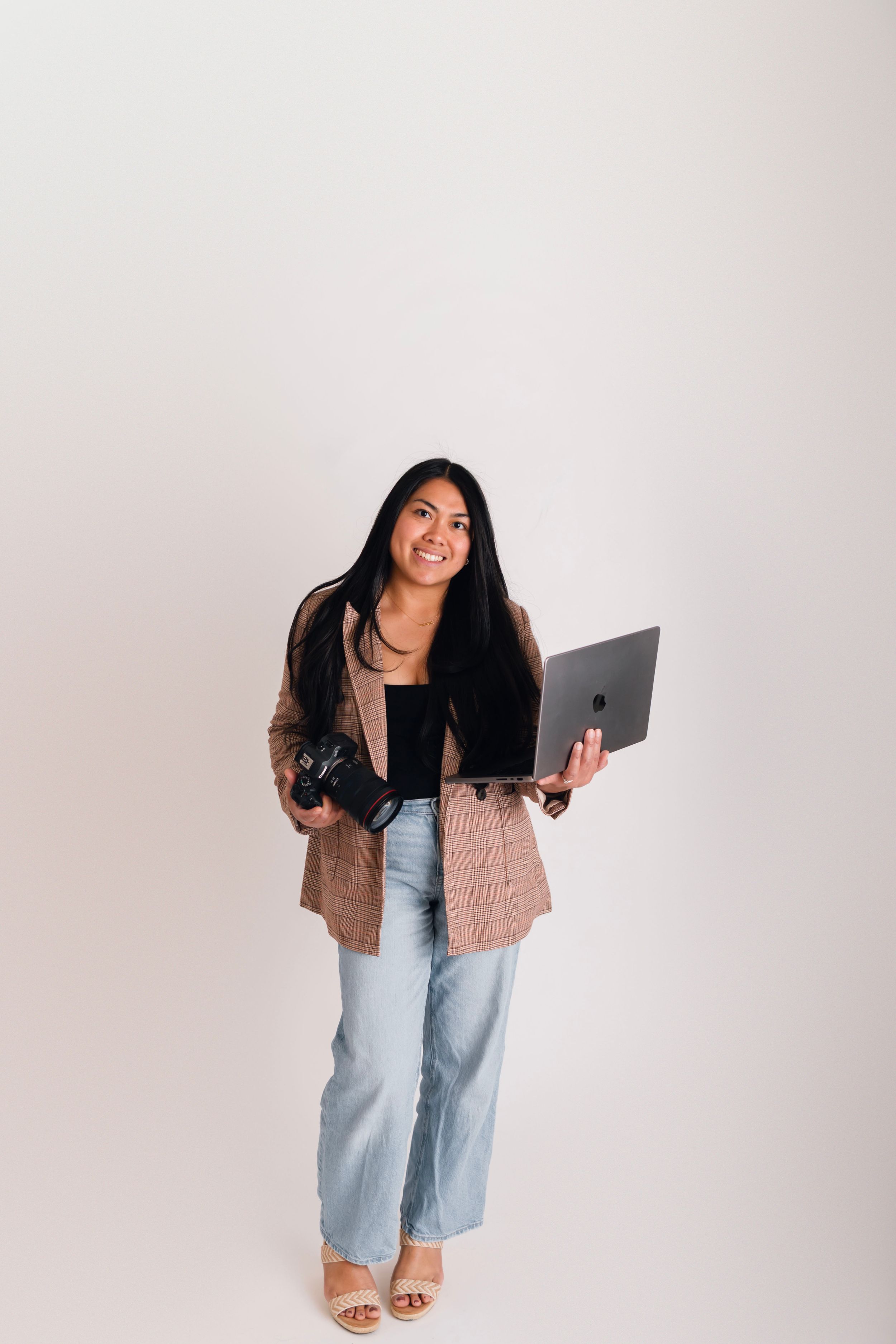 Smiling woman holding a camera and laptop, wearing a blazer and jeans, standing against a white background.