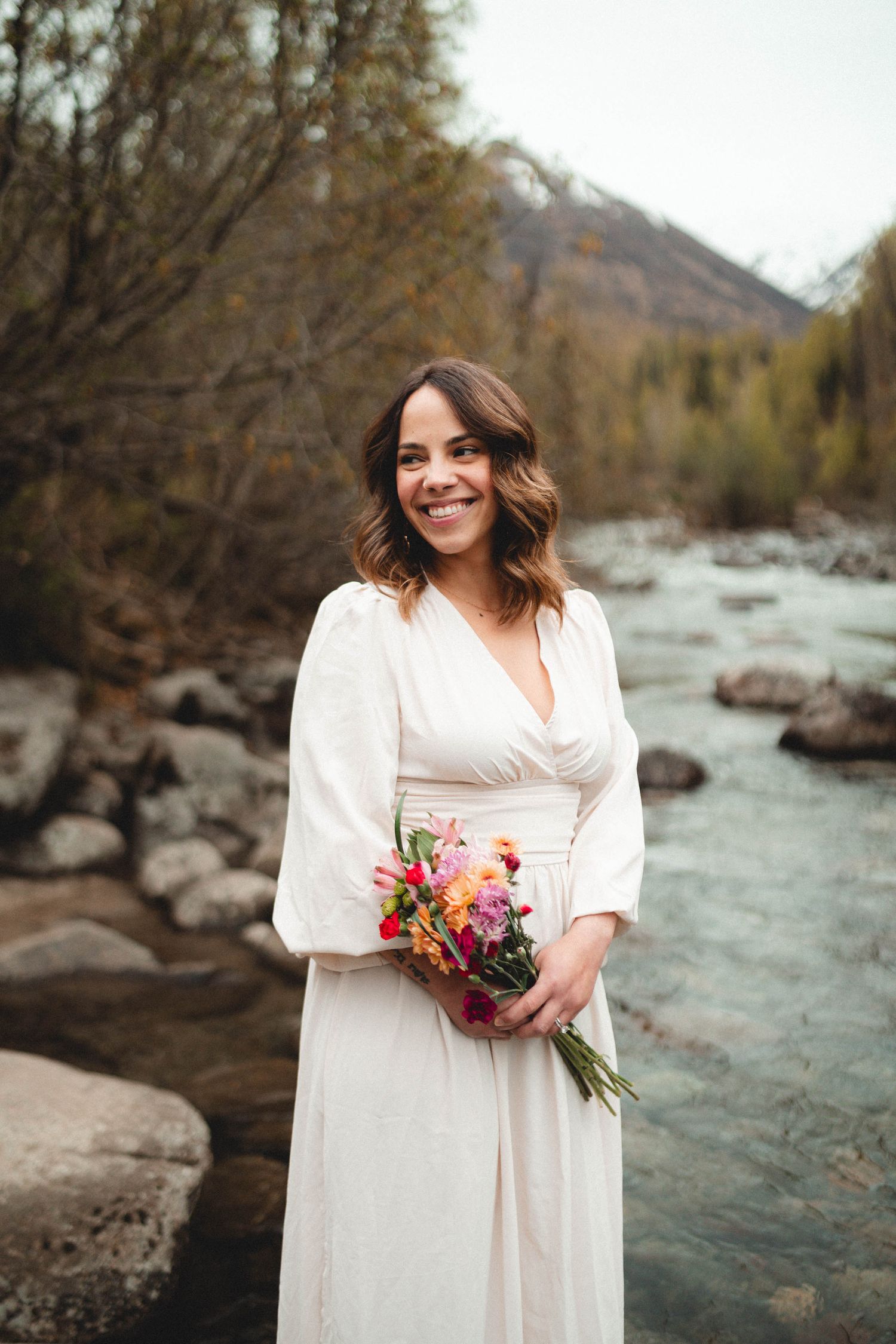 Smiling bride in white dress holding colorful bouquet standing by rocky mountain river with autumn trees.