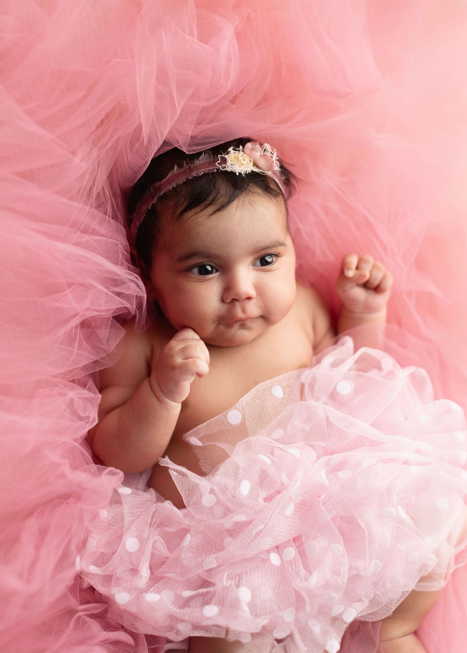 Adorable baby girl in pink tutu skirt and floral headband lying on fluffy pink tulle backdrop.
