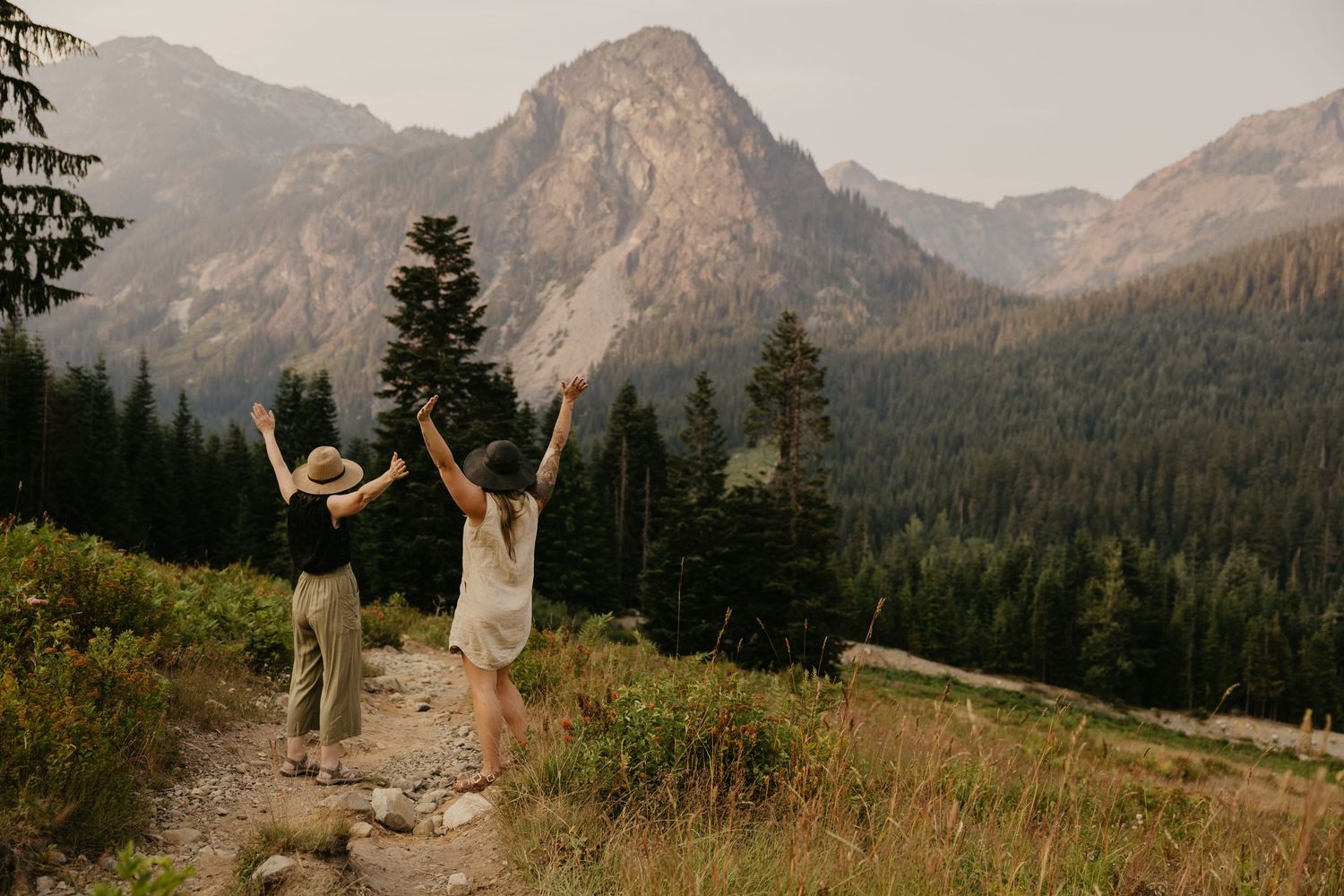 Couple with arms raised celebrating on a mountain trail surrounded by tall pine trees and dramatic peaks in the pnw during golden hour