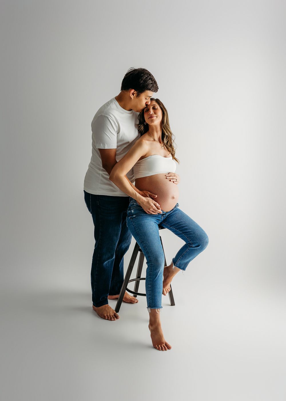 A couple poses for a maternity photo, the man kissing her head while she cradles her baby bump in a studio.