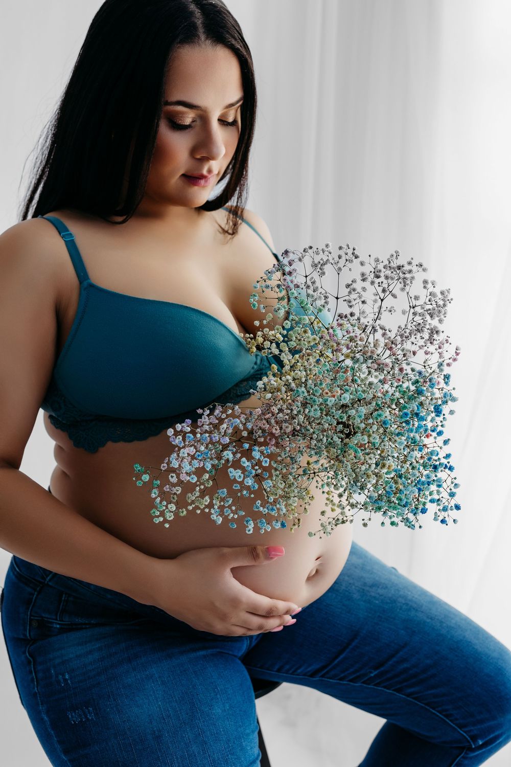 Pregnant woman in teal bra and jeans holding colorful wildflowers over her baby bump in a bright studio.