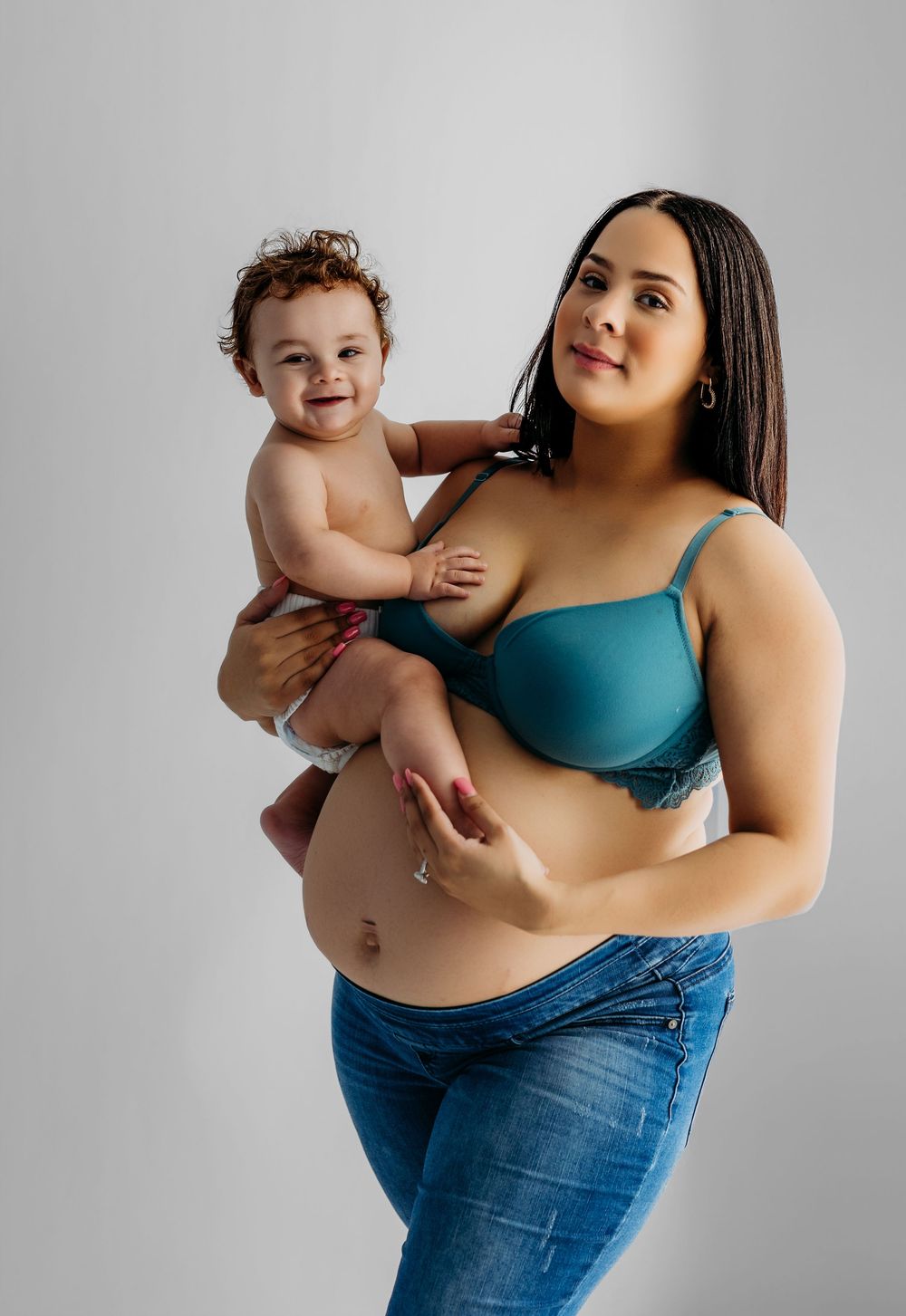 A young mother in a teal bra and jeans holds her smiling baby in a studio portrait photo.