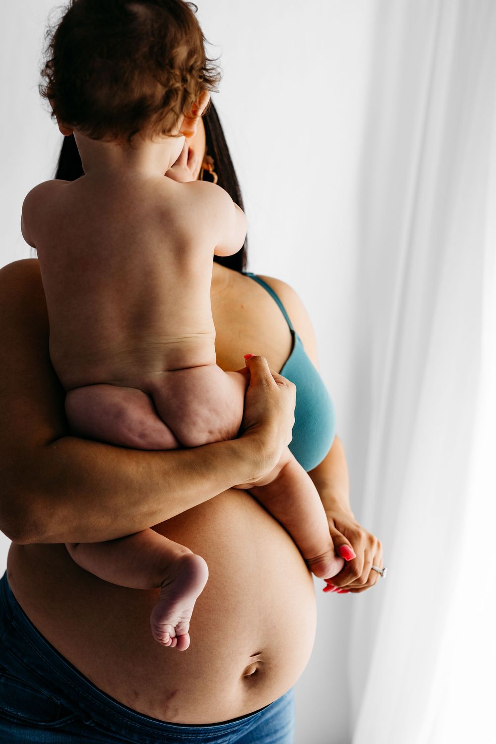 A mother in a teal bra holds her chubby baby against her pregnant belly near a bright window.