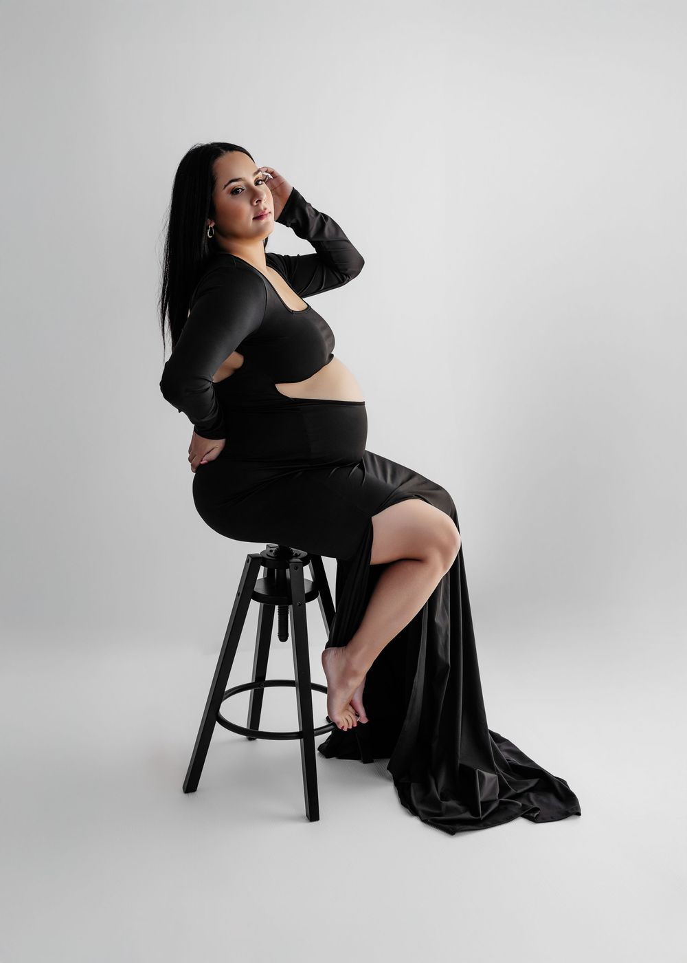 Pregnant woman in black crop top and maxi skirt seated on stool, maternity photography studio portrait.