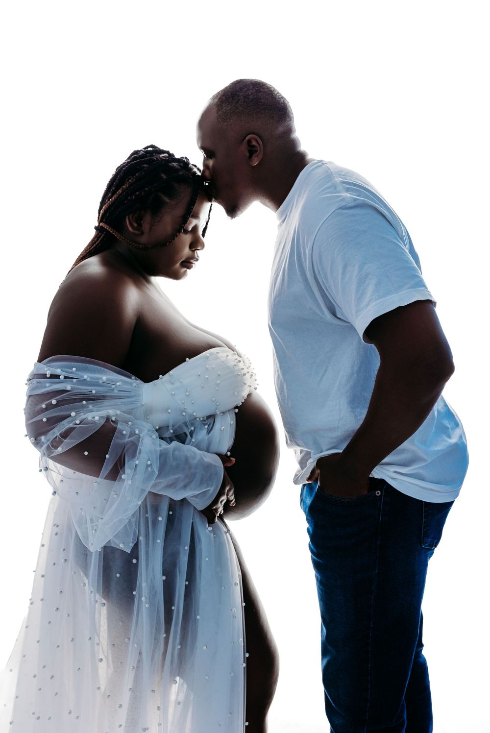 Pregnant couple in maternity photoshoot, woman draped in white tulle fabric, man in blue shirt, silhouetted against white background.