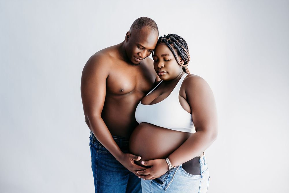 A shirtless man embraces a pregnant woman in a white crop top and jeans during a maternity photo shoot.