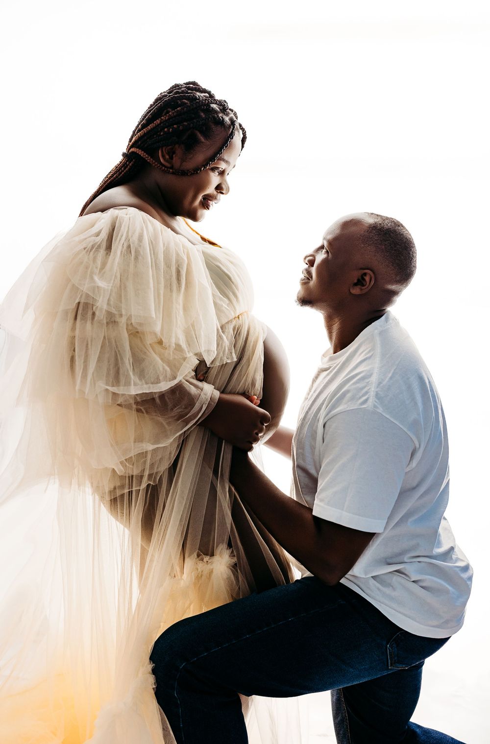 A man kneels before a pregnant woman in a sheer gown during a maternity photo shoot.