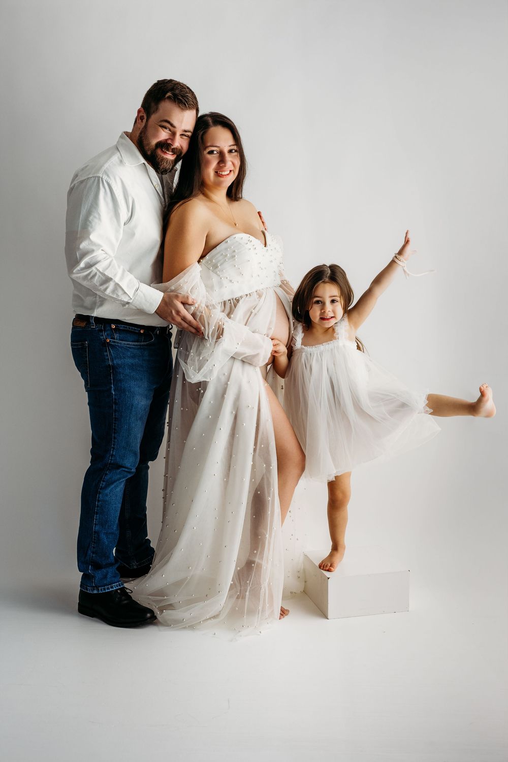 Happy family maternity photo with pregnant woman in white gown, man in jeans, and excited little girl on white background.