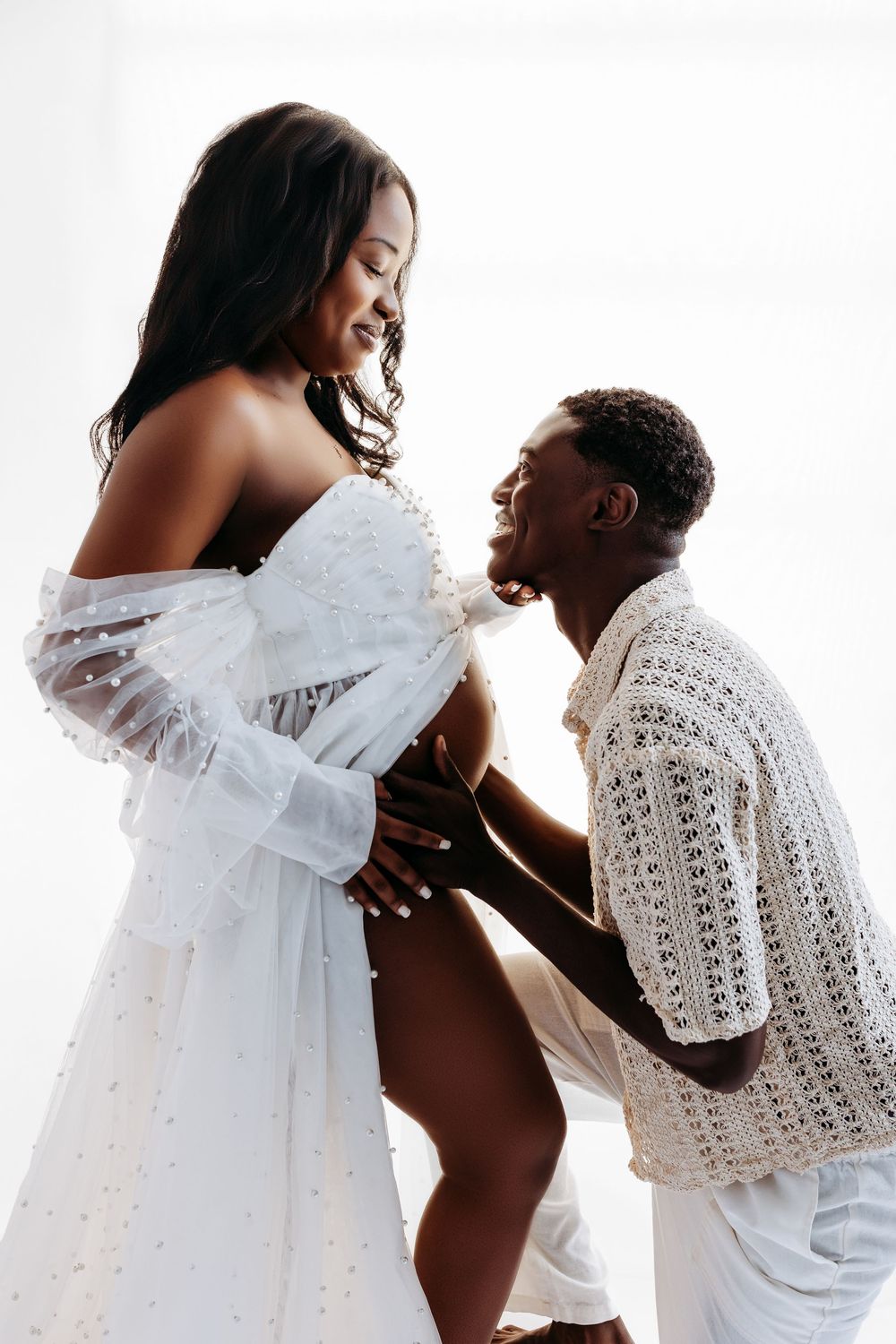 A joyful couple in white celebrating pregnancy, with the man tenderly kissing the woman's baby bump.