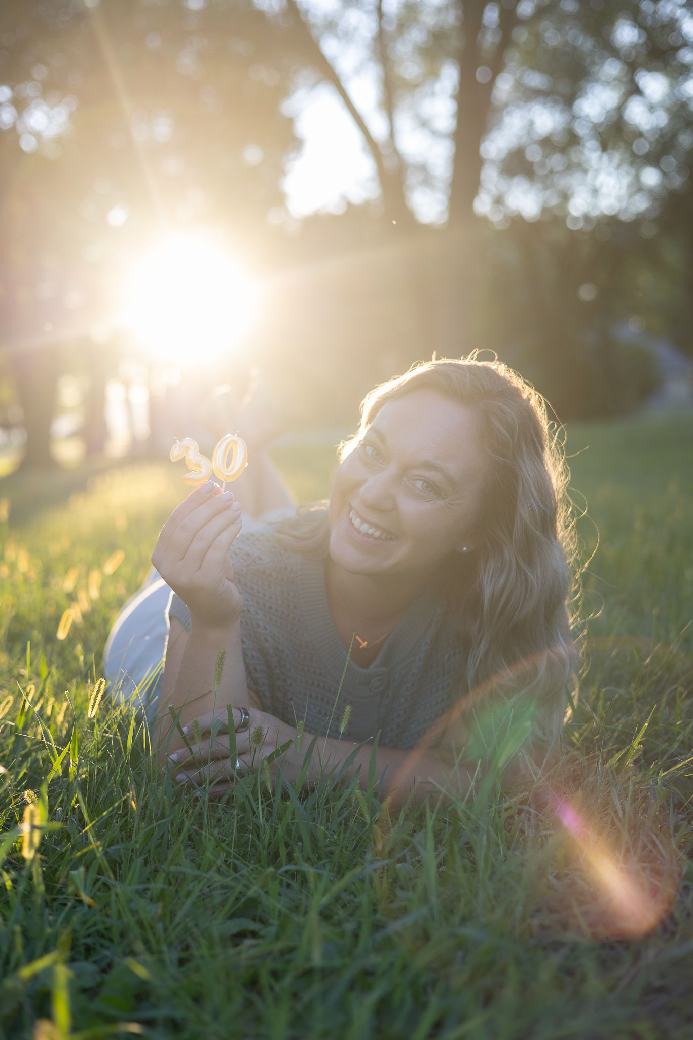Joyful woman lying on grass holding a flower with sunlight streaming through trees behind her.