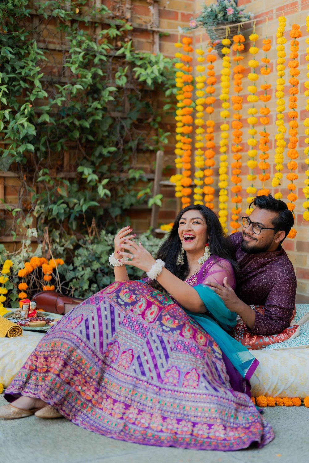 Couple in vibrant Indian attire taking a selfie, seated before marigold garland backdrop at a festive celebration.