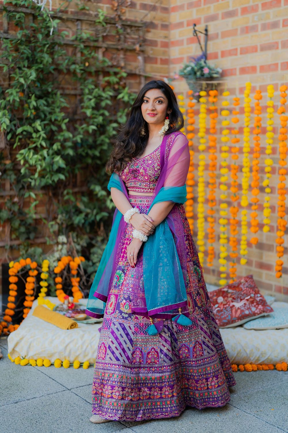 Woman in vibrant purple embroidered lehenga choli with teal dupatta poses at Indian celebration with marigold garlands.