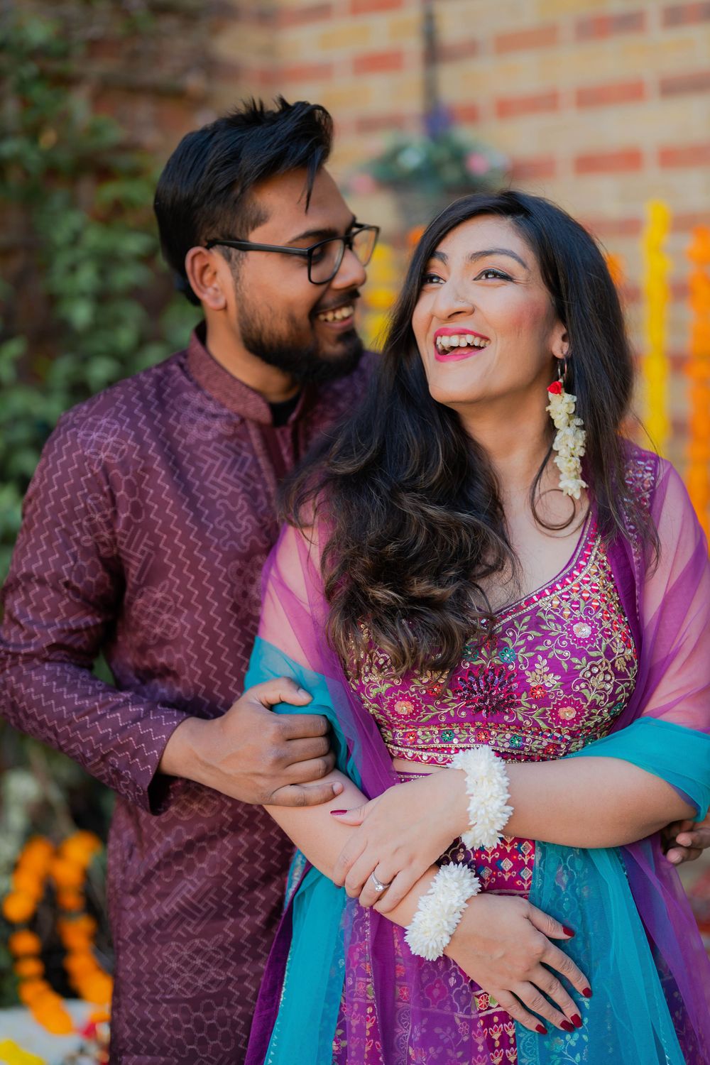 A joyful Indian couple in traditional attire embrace outdoors, the woman in blue and pink lehenga, man in maroon kurta.