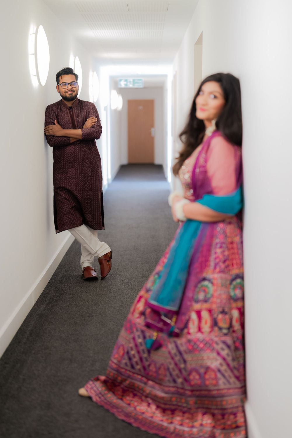 Couple in traditional South Asian attire posing in a hallway, man in dark kurta, woman in vibrant pink lehenga.