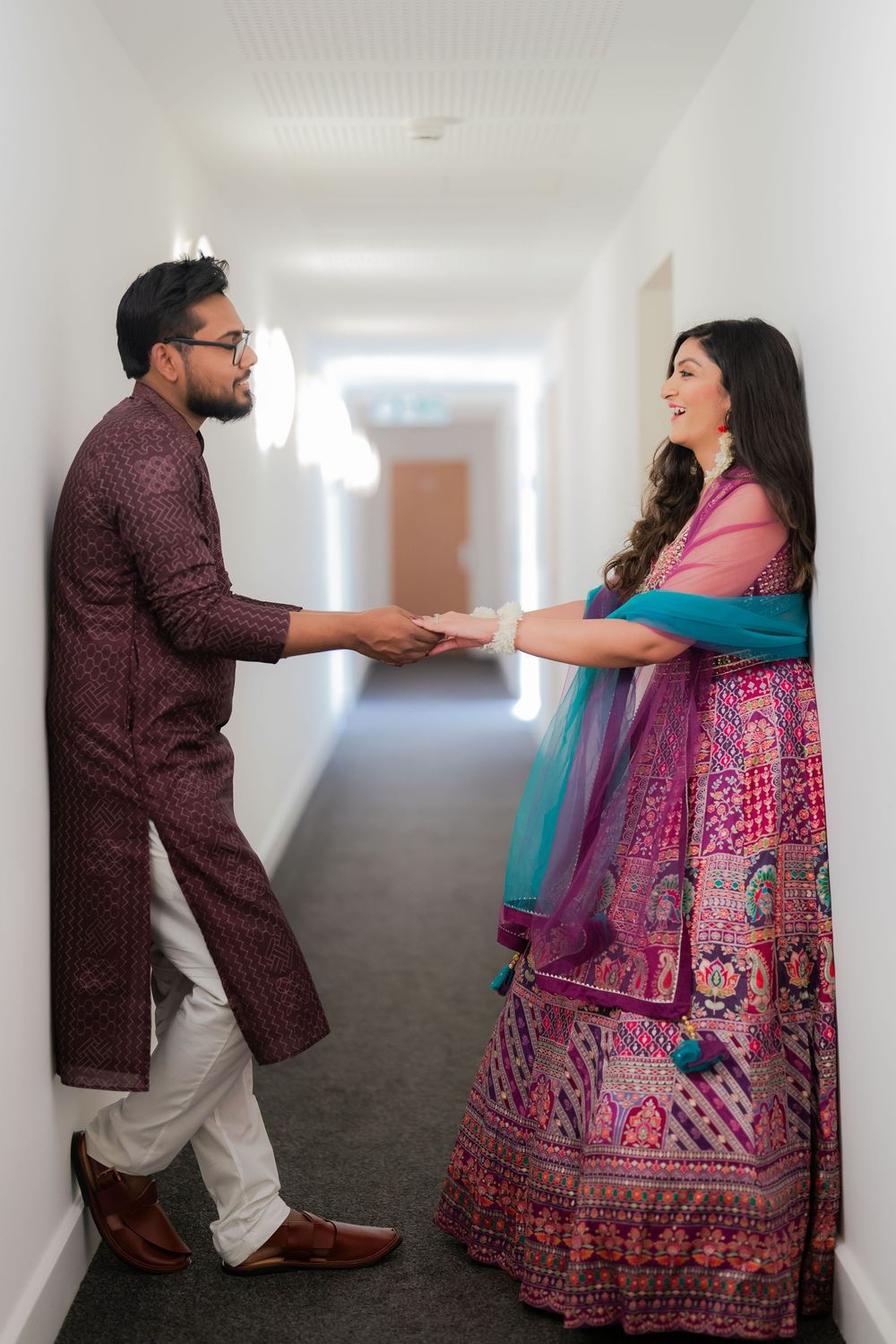 Indian couple in traditional attire holding hands in a hallway, woman in pink lehenga, man in maroon kurta.