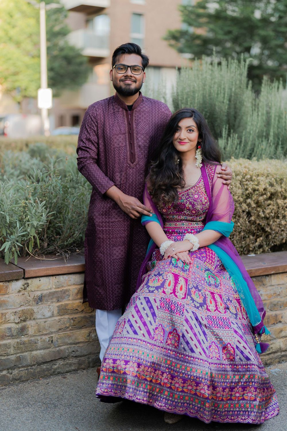 Couple in coordinating purple Indian traditional attire posing outdoors, woman in embroidered lehenga, man in sherwani.