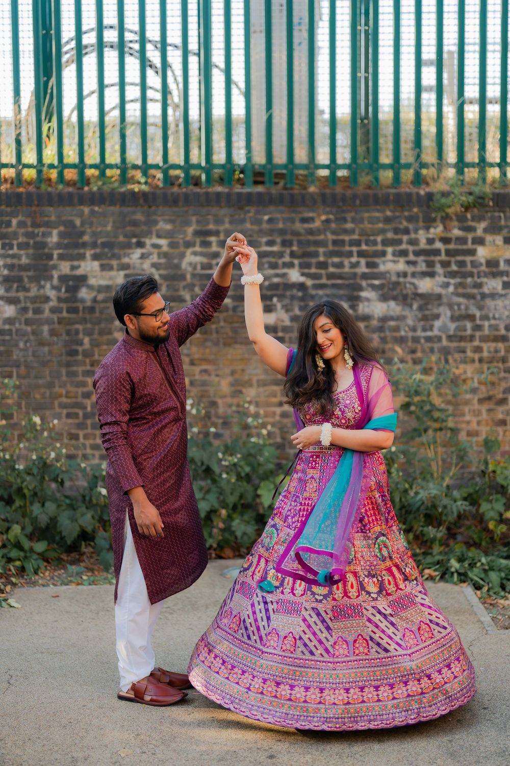 Couple in traditional Indian attire dancing outdoors, woman in vibrant pink lehenga, man in maroon kurta, brick wall backdrop.
